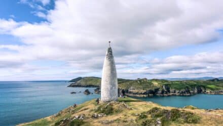A photograph capturing the Baltimore Beacon in County Cork, Ireland. The image showcases the landmark against a coastal backdrop. The beacon is a simple structure characterized by its vertical white form. The coastal scenery features cliffs and the expanse of the Atlantic Ocean.