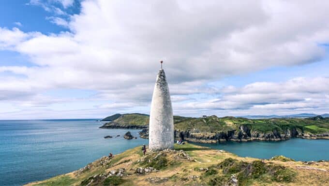 A photograph capturing the Baltimore Beacon in County Cork, Ireland. The image showcases the landmark against a coastal backdrop. The beacon is a simple structure characterized by its vertical white form. The coastal scenery features cliffs and the expanse of the Atlantic Ocean.