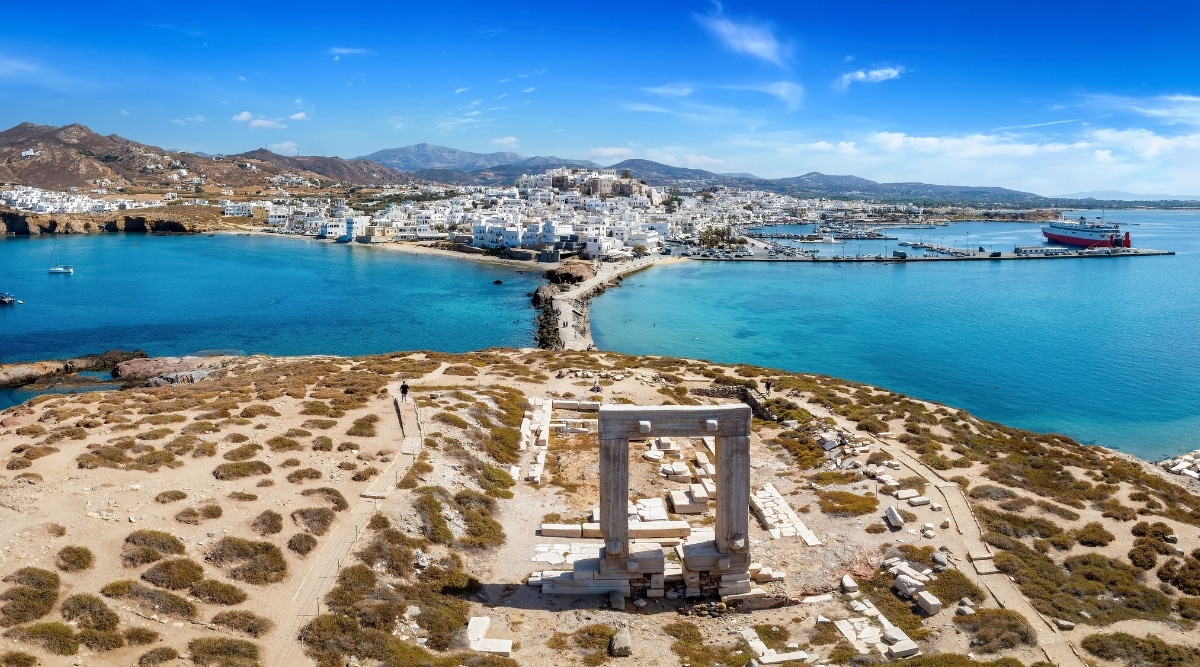 An aerial view capturing the renowned Portara Gate on Naxos island in the Cyclades, Greece. The image features the ancient stone structure set against the backdrop of the town and harbor. The Portara Gate, a prominent historical landmark, is visible with its distinctive architectural details.