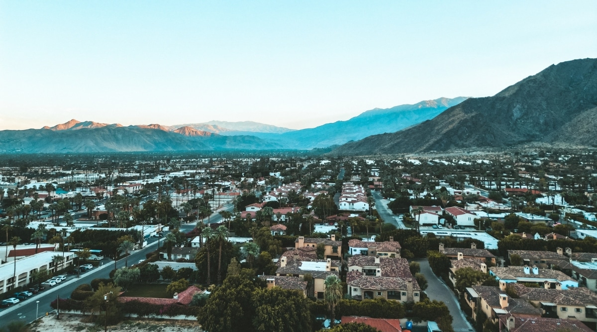 An aerial photograph capturing Palm Springs, California, at dawn. The clear sky above the city provides a serene backdrop, while the one-story buildings below contribute to the city's characteristic architectural profile. In the background, mountain ranges and hills add to the natural topography of the region. The image features abundant greenery throughout the city.