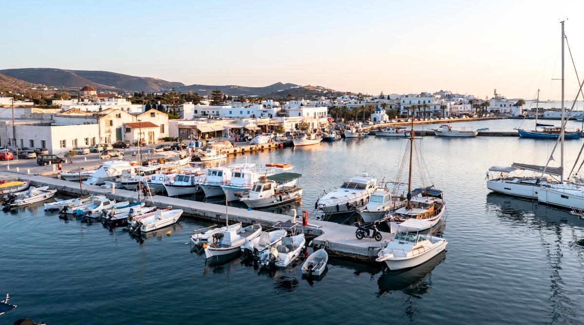 Aerial drone view of Parikia port marina in the afternoon on Paros Island, part of the Cyclades in Greece. The image depicts yachts, sailboats, and fishing boats moored at the harbor dock.