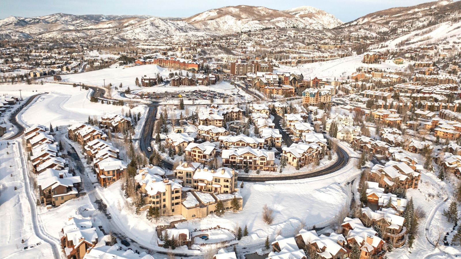 An aerial perspective capturing the snowy cityscape of Steamboat Springs. The image depicts a vast expanse of snow covering houses, rooftops, and the surrounding landscape. Numerous houses are visible within the snowy terrain. The hills in the background are also covered with snow.