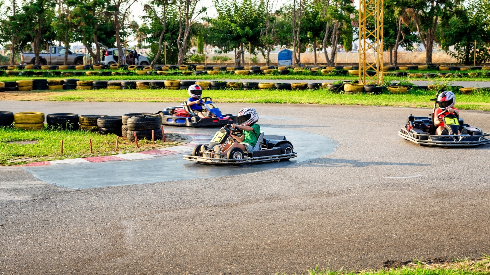 A photograph depicting three children in a go-kart driving. The children are wearing helmets for safety. The go-kart road is marked by rubber tires from the go-kart, placed along the route.