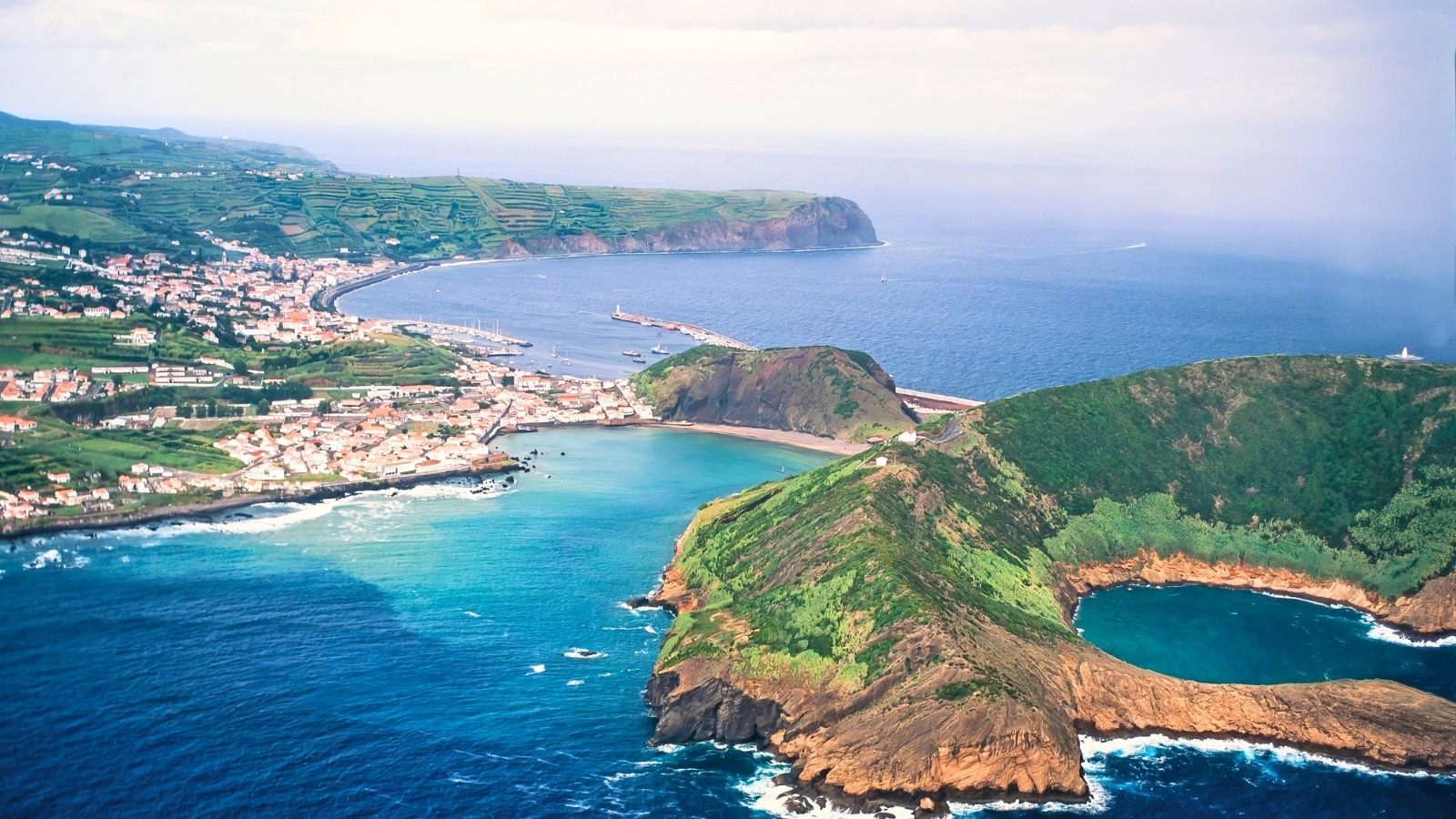 A top view photograph capturing the cityscape of Azores, Portugal. The image features mountains in the foreground, situated along the water, with a body of water in between. The city is visible in the background.