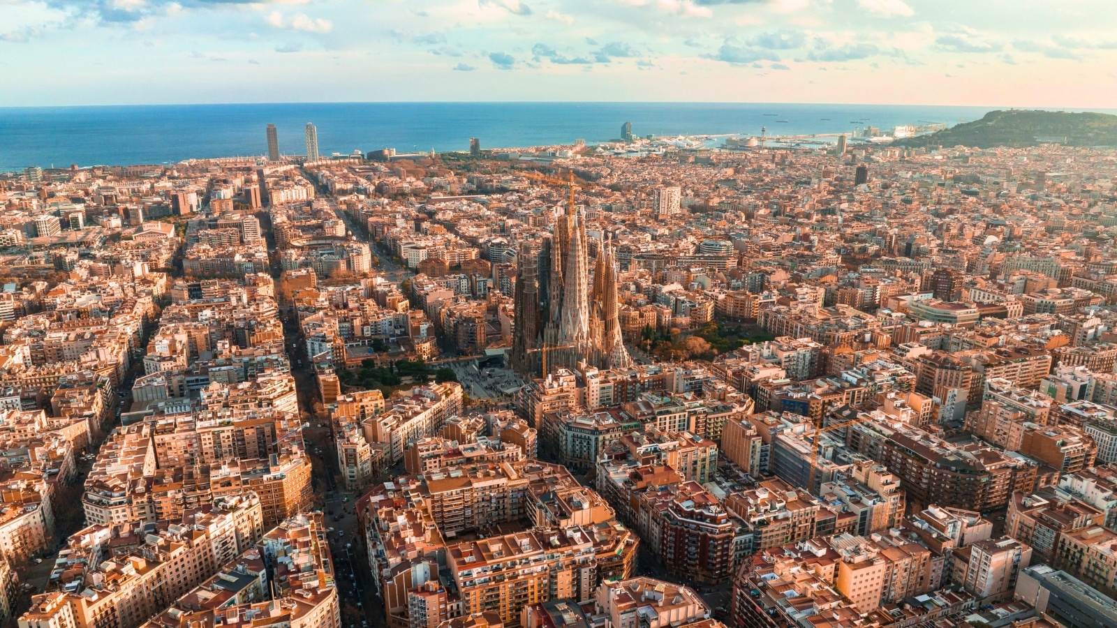 An aerial view capturing the Barcelona City skyline at sunset, featuring the iconic Sagrada Familia Cathedral. The image showcases the Eixample residential area known for its famous urban grid, characterized by typical urban octagon blocks.
