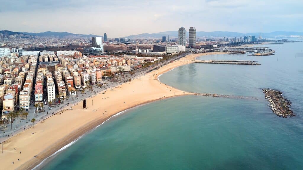 A view of Barcelona Beach with the cityscape of Barcelona in the background. The image depicts a sandy beach with a calm sea under cloudy weather conditions. The vast urban expanse of Barcelona serves as a prominent backdrop.