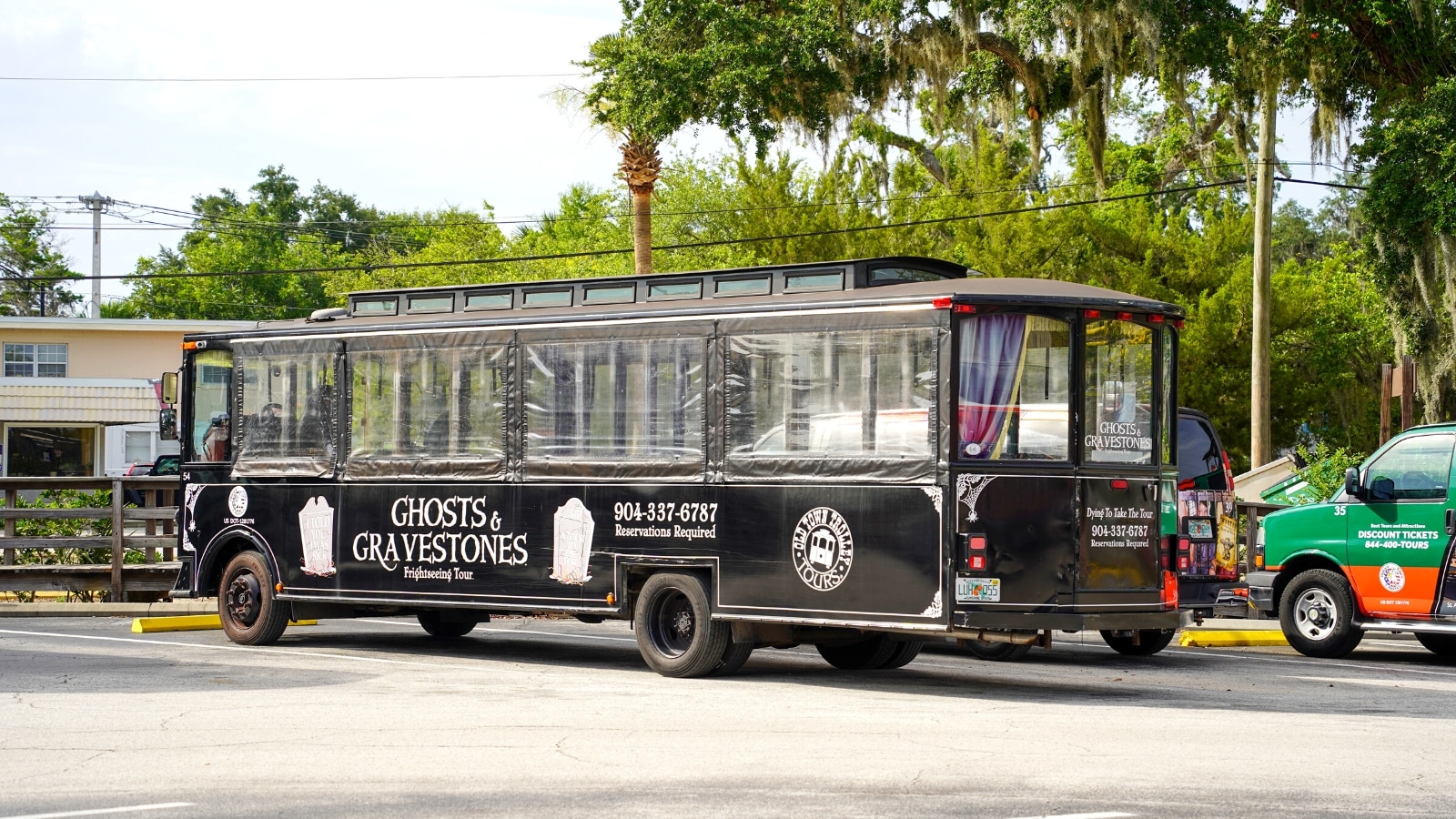 A photograph featuring a black bus prominently labeled 'Ghost' with gravestones in the background. The image captures the exterior of the Ghost Tours of St. Augustine vehicle, showcasing the branding and theme associated with the tour service. 