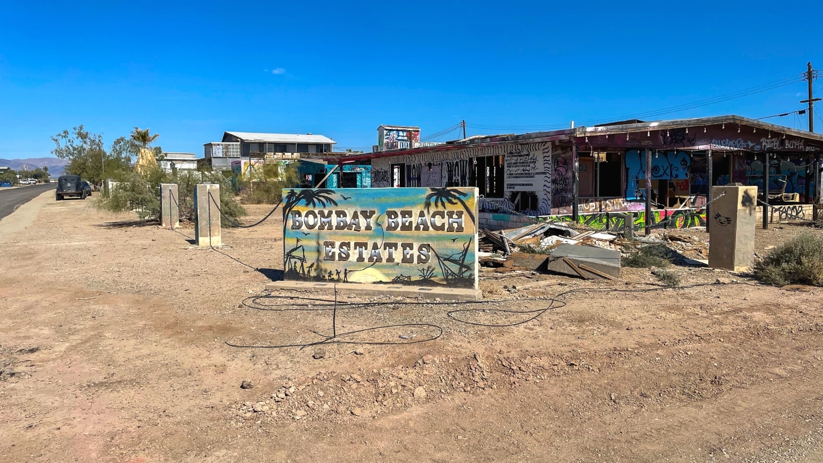 A photograph depicting buildings at Bombay Beach, California, featuring artistic designs. The image includes a view of the Bombay Beach Estates sign. The buildings exhibit creative elements, contributing to an artistic ambiance within the area.