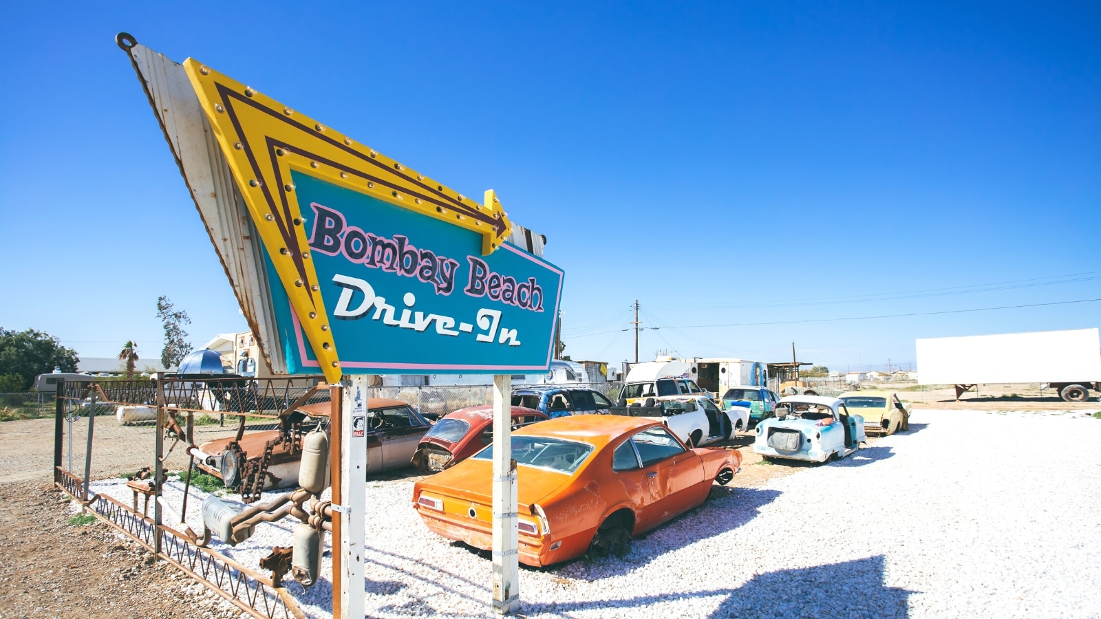 The photograph depicts the Bombay Beach Drive Inn, featuring a foreground sign bearing the establishment's name. The sign is set against a backdrop of a parking lot containing various old cars of diverse colors and brands. Many of these cars lack wheels, and some are observed without doors. The sky is characterized by a clear, pure blue hue.