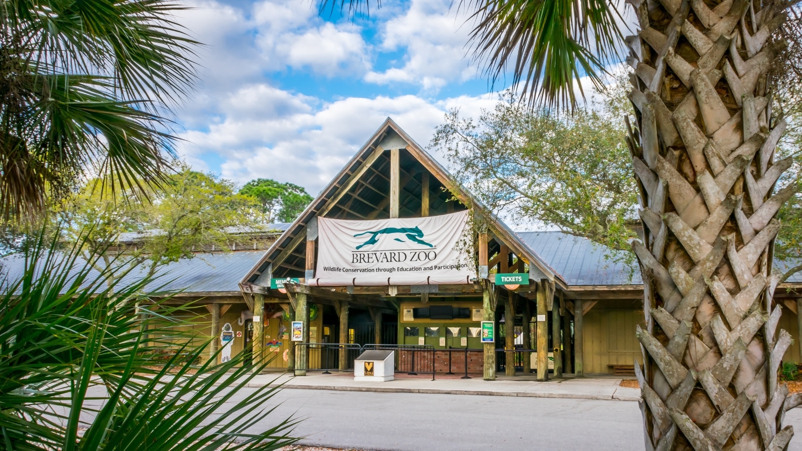 A view of the Brevard Zoo entrance without any individuals present. The sky above features white clouds, creating a neutral atmospheric backdrop. The entrance is characterized by a triangular roof structure, and at the center is a sign prominently displaying the name 'Brevard Zoo.'