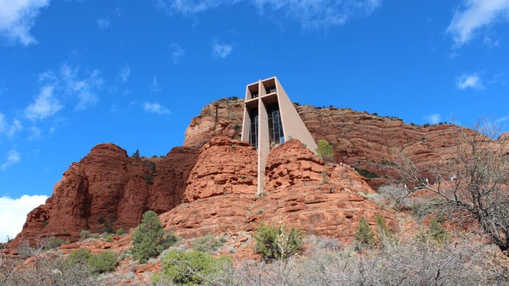 A photograph capturing the Chapel of the Holy Cross in Sedona, Arizona. The image showcases the chapel's distinctive architecture against the backdrop of the surrounding red rock formations. The chapel appears as a focal point in the landscape, with minimal vegetation and clear skies.