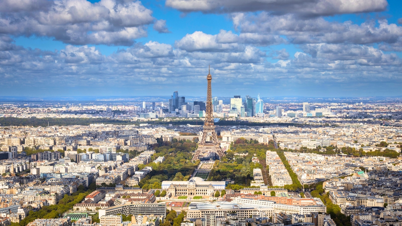 A cityscape photograph depicting Paris, France, on a sunny day with the iconic Eiffel Tower prominently visible. The image captures the architectural charm of Paris, featuring buildings, streets, and landmarks typical of the city's urban landscape.