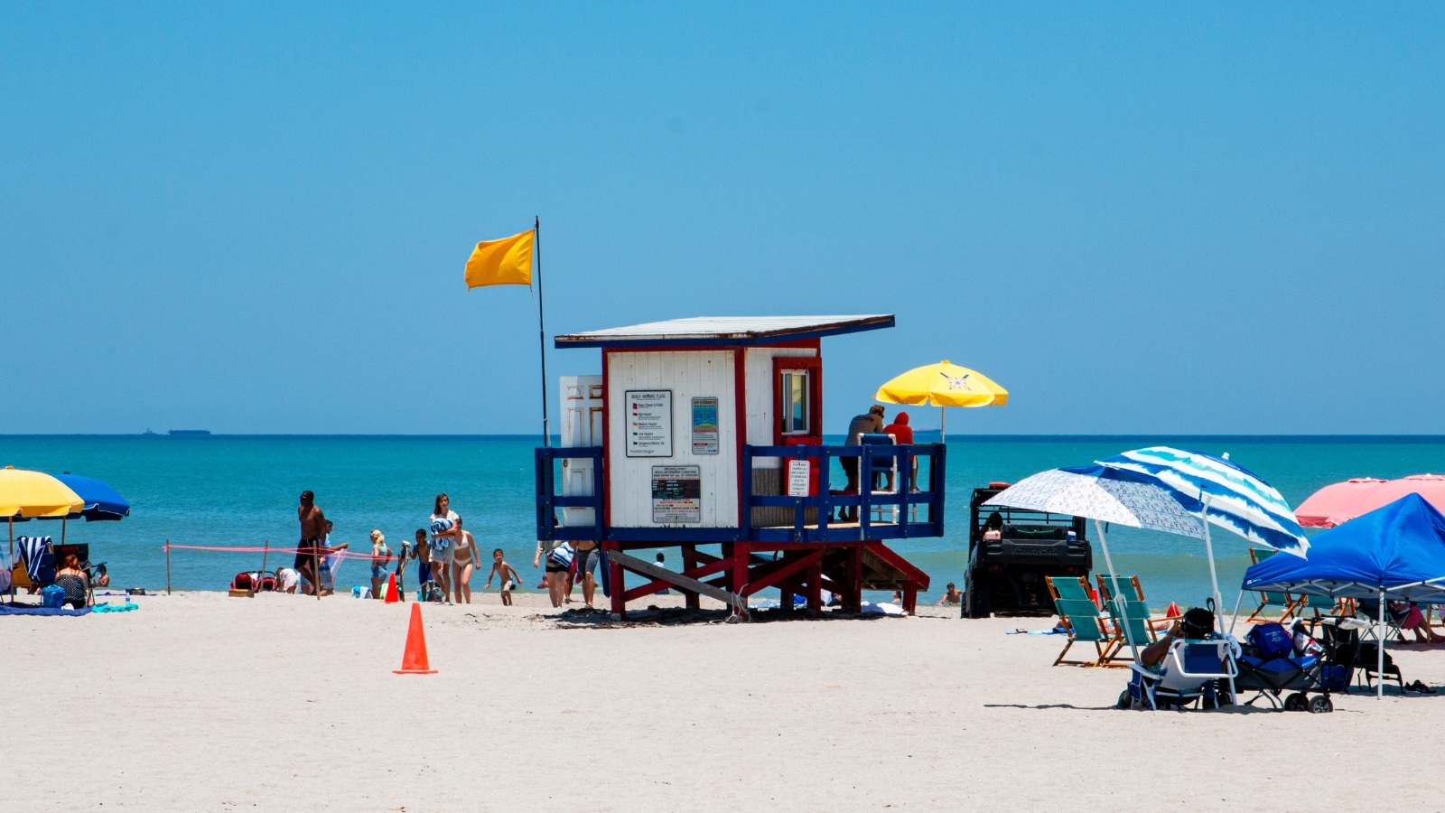 A beach view capturing the scene at Cocoa Beach, Florida. In the center of the image stands a rescue booth with a yellow flag. Numerous visitors surround the beach booth, accompanied by sun umbrellas of various colors. The sky overhead is clear blue, and the ocean presents a calm disposition.