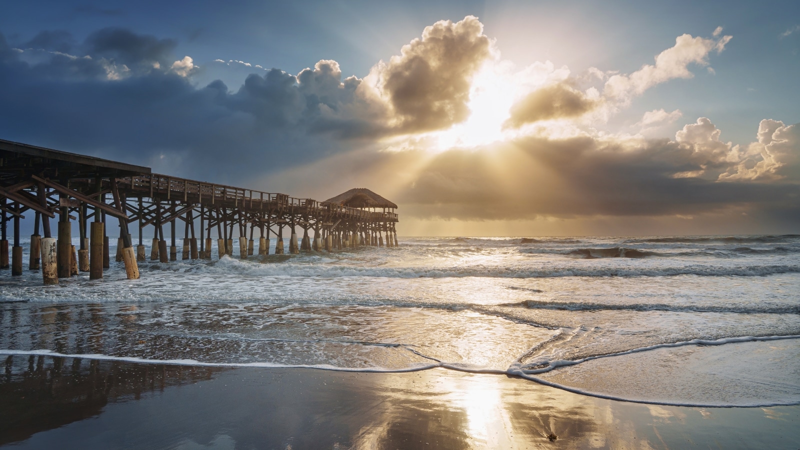 A photograph capturing Cocoa Beach Pier from the shoreline. The image depicts the pier extending into the ocean under a dramatic sky. Sunlight breaks through large dark clouds, creating a striking visual effect.