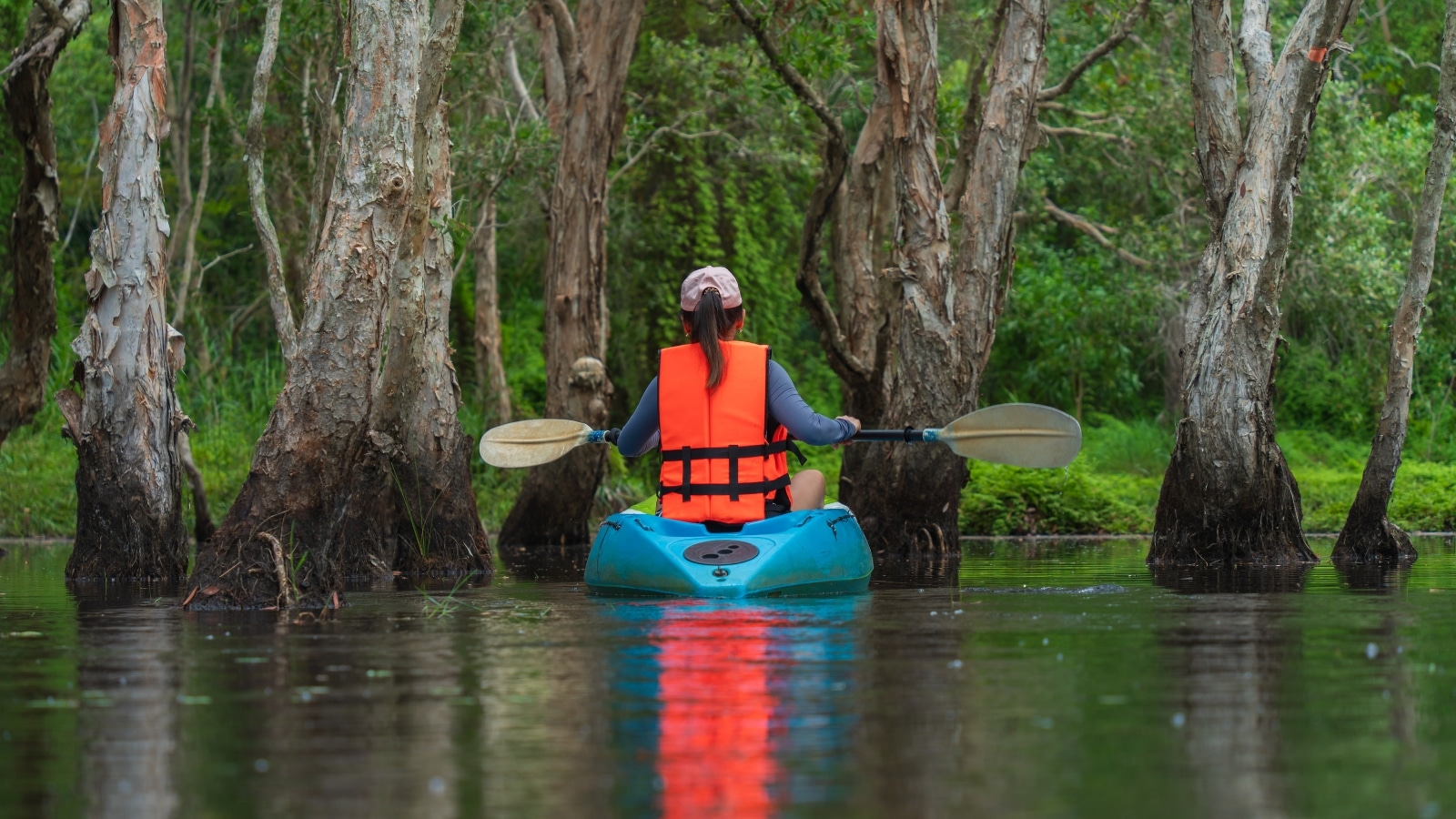A photograph depicting a girl kayaking on a blue kayak wearing an orange vest and a pink cap. The scene is surrounded by greenery and trees.