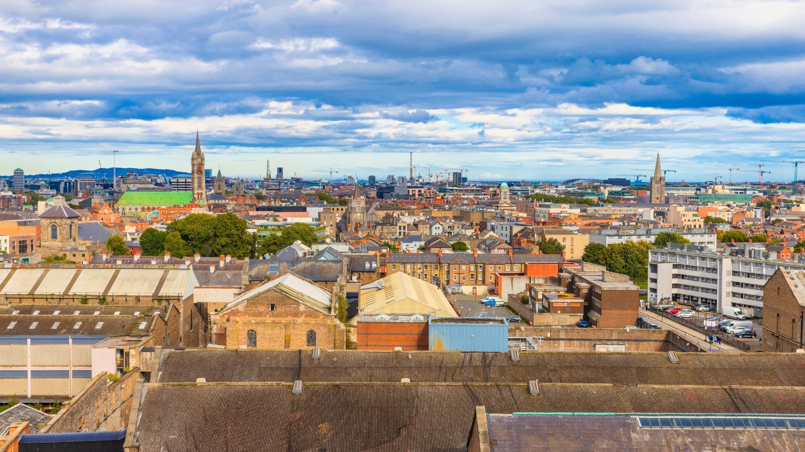 An aerial photograph capturing the Dublin skyline on a cloudy day. The image presents a comprehensive view of the cityscape, showcasing various architectural structures and urban developments. The overcast weather conditions are evident with clouds covering the sky.