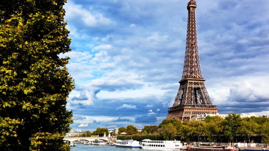 A photograph showcasing a unique perspective of the Eiffel Tower and Seine River in Paris, France. The image is framed by trees, providing a natural element to the composition. Tourist boats can be seen navigating the river