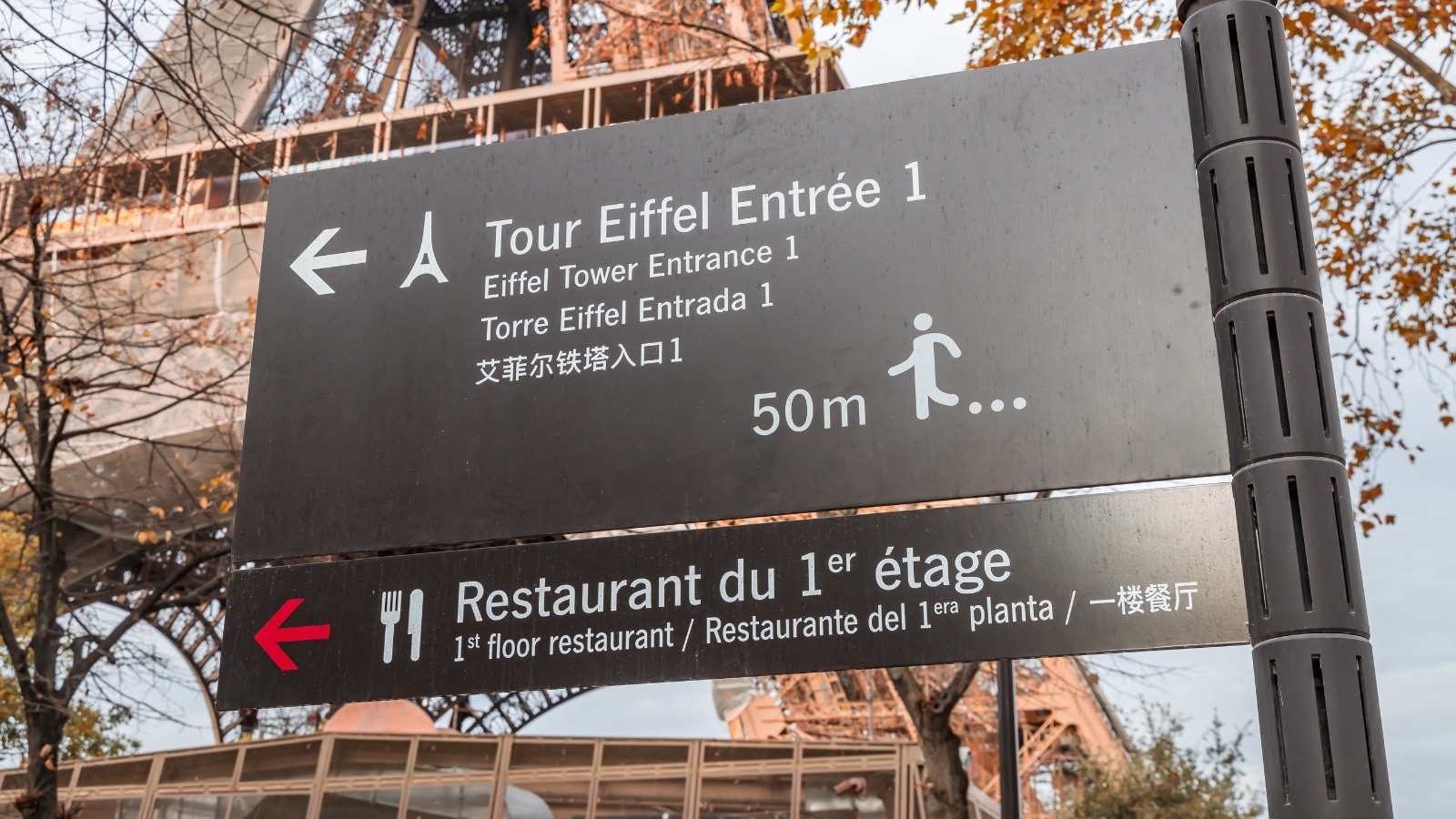 A close-up photograph capturing the sign near Entrance 1 of the Eiffel Tower, Paris. The image includes a sign indicating the presence of a restaurant on the first floor of the iconic landmark.