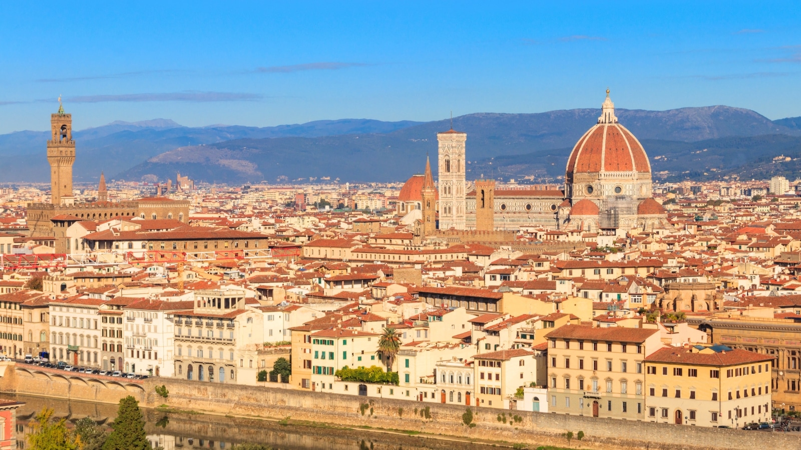 A panoramic photograph capturing the cityscape of Florence from Michelangelo Hill in Tuscany, Italy. The image offers a wide view of the urban layout, highlighting key architectural landmarks and the Tuscan landscape.