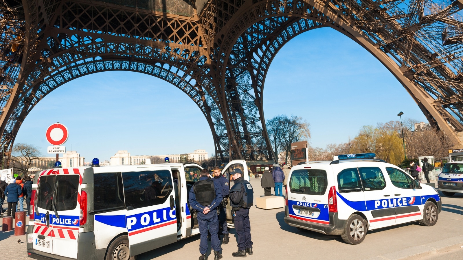 A photograph depicting French police guards stationed near the Eiffel Tower in Paris. The image includes two police cars and several police officers in the vicinity.