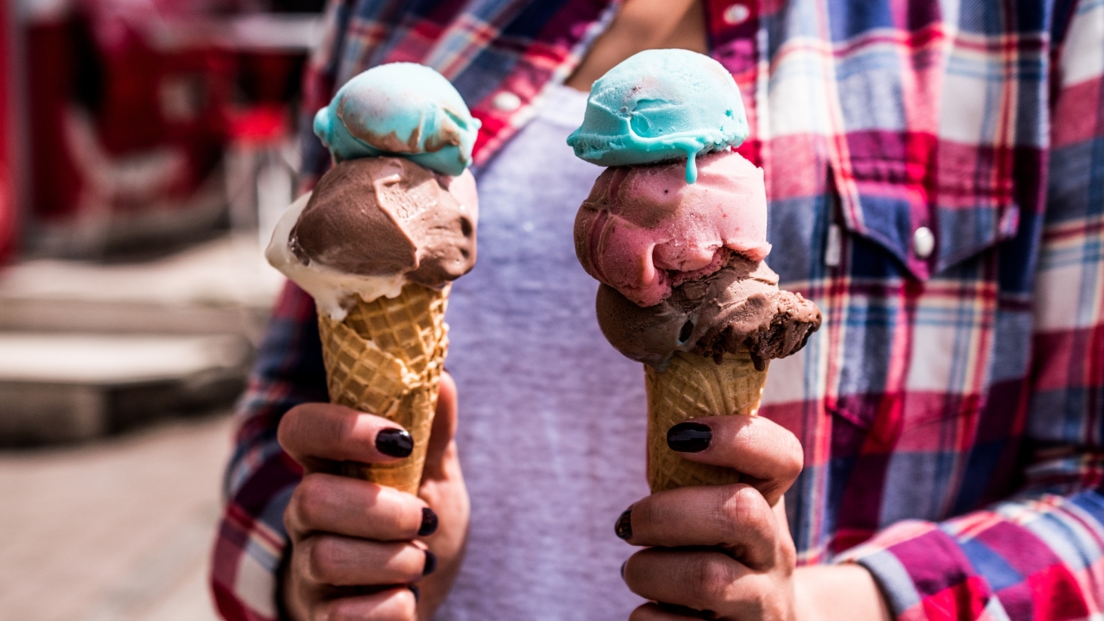 A photograph depicting a girl in a plaid shirt holding two ice cream cones. One cone features three scoops of ice cream, while the other has two.