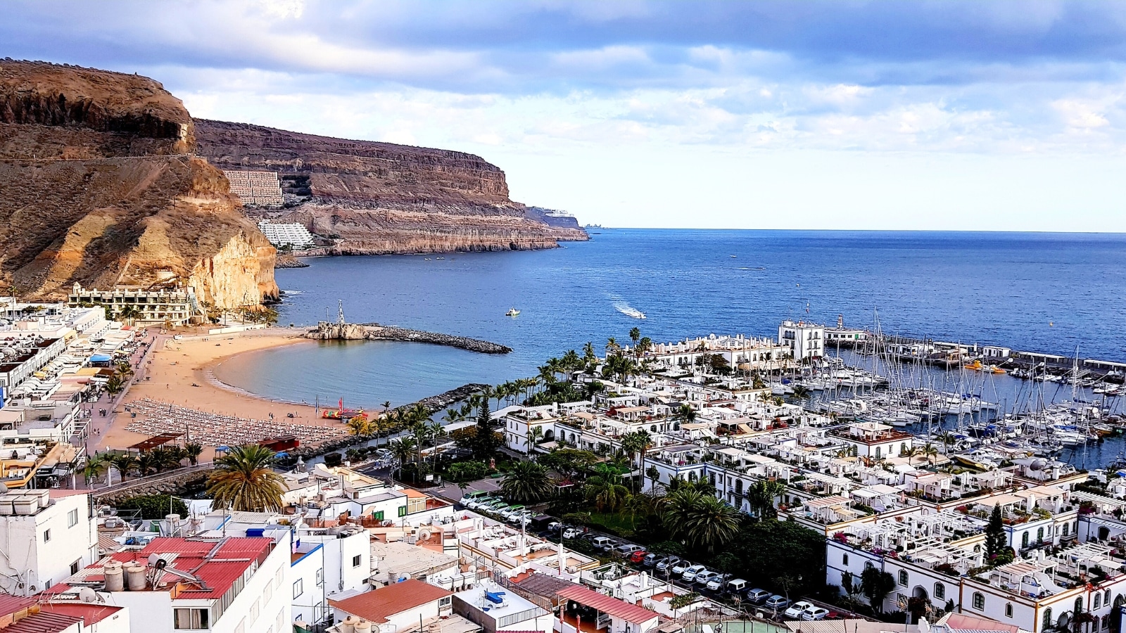 A scenic photograph capturing a view of a city near the ocean and mountains on Gran Canaria, an island in the Canary Islands. The image features a landscape with a combination of urban structures, the coastline, and mountainous terrain.