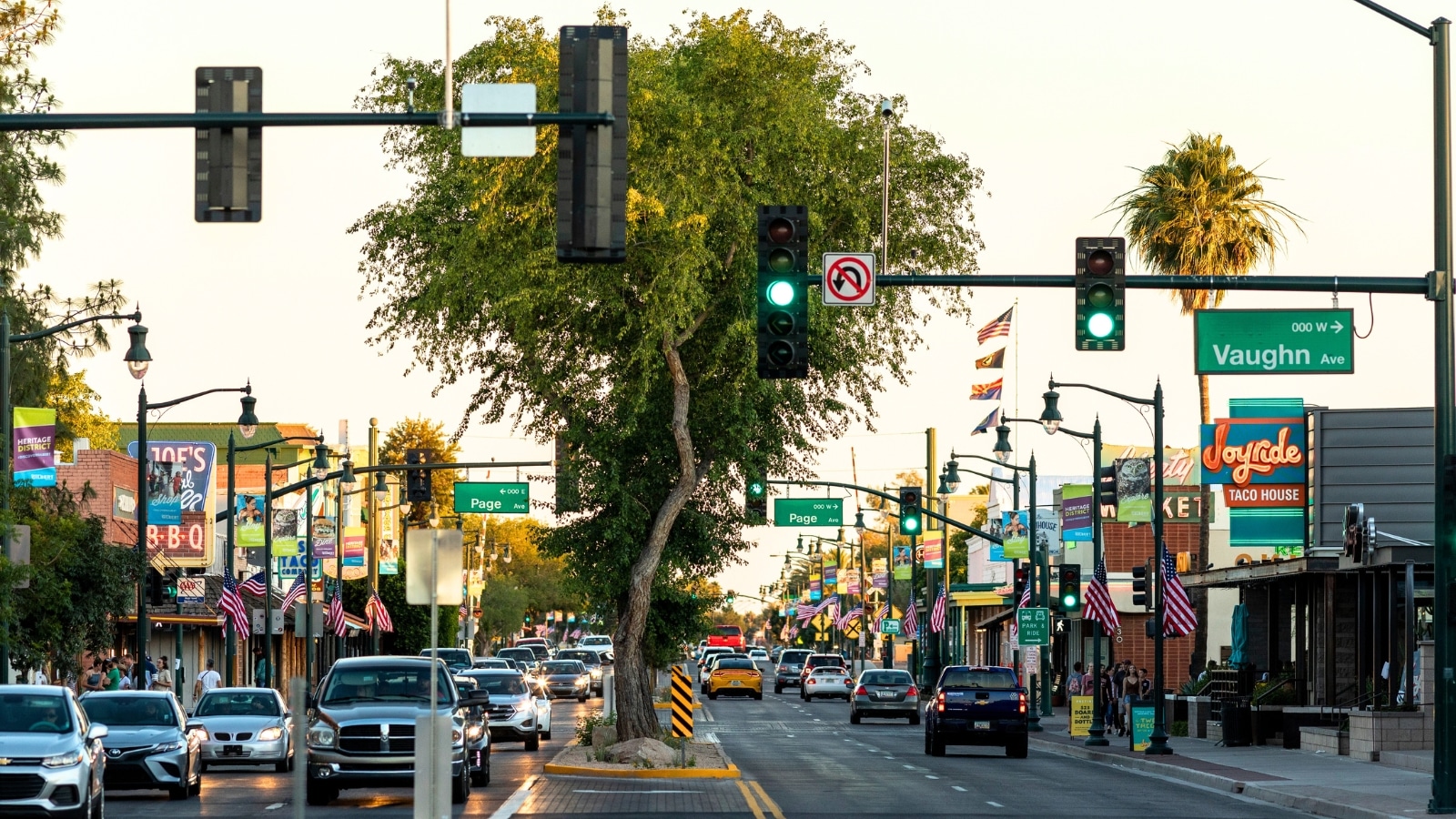 A photograph depicting a bustling scene along Main Street in Gilbert, Arizona. The image showcases a multitude of cars traveling along the road, flanked by buildings and trees lining the street. 