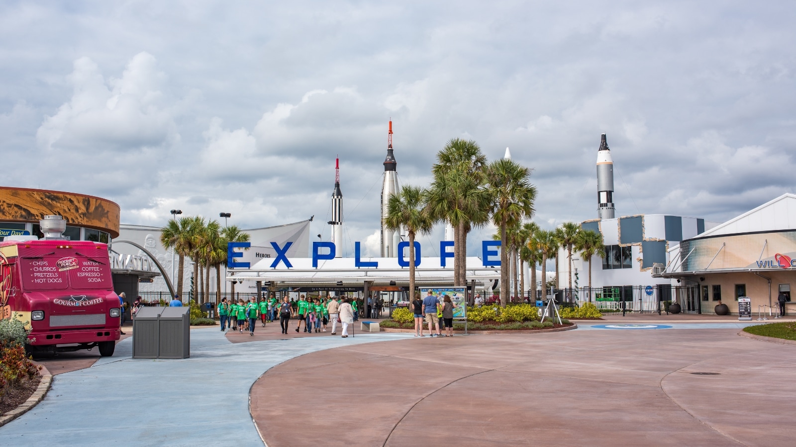 A view of the entrance to Kennedy Space Center with prominent signage displaying the word 'Explore' in large letters. The entrance is marked with a straightforward and functional design. In the background, several rockets are visible, contributing to the space-themed atmosphere.