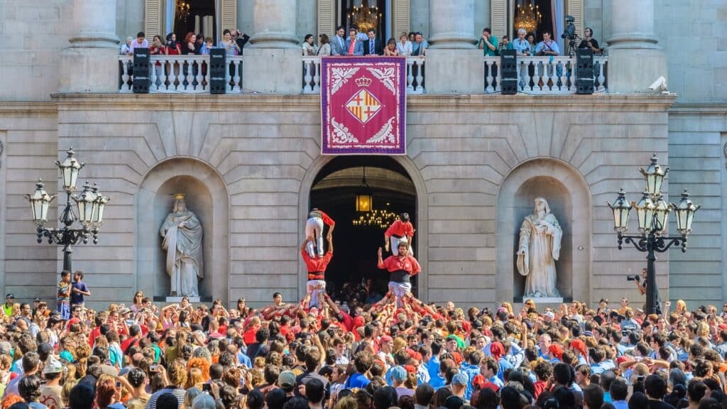 A photograph capturing the La Mercè annual festival in Barcelona. The image features a large crowd gathered in front of a building, with two distinct groups wearing red clothes engaged in a climbing activity.