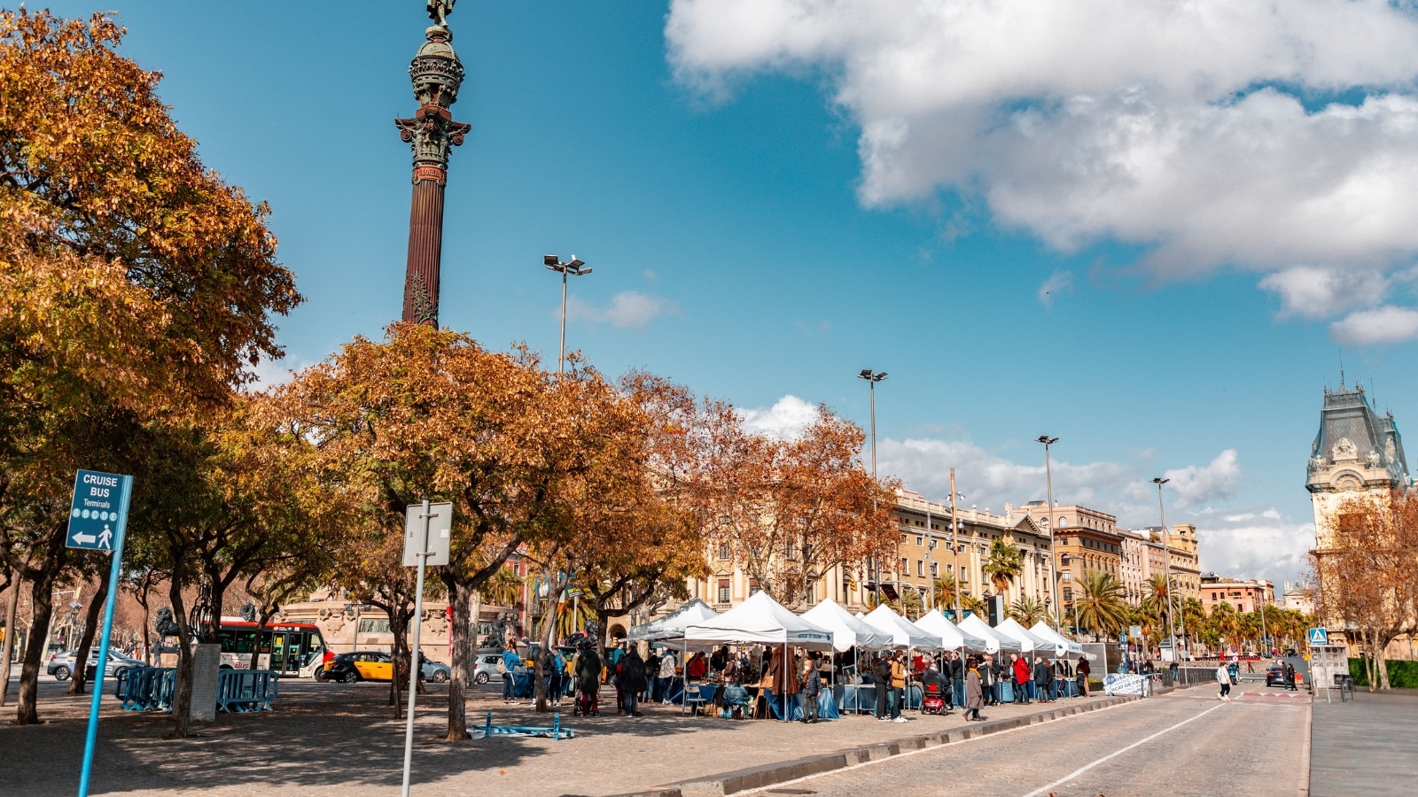 A photograph capturing Placa Portal de la Pau, a busy city square located at the lower end of La Rambla in Barcelona, Catalonia, Spain. The image depicts the bustling urban environment with pedestrians and activities in the square.