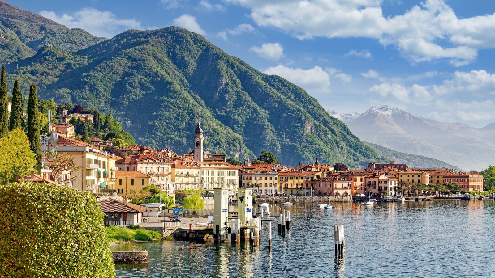 A photograph depicting Menaggio, a town situated on Lake Como in the Italian Lake District. The image captures the lakeside setting with surrounding hills and buildings. Lake Como is visible, showcasing its calm waters and mountainous backdrop.