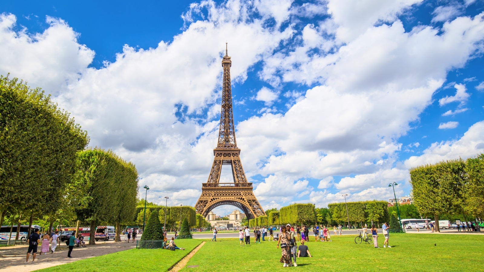 A panoramic view capturing the Eiffel Tower from the gardens in front of the tower. The image features abundant greenery and trees, with people scattered along the lawn. The Eiffel Tower stands prominently in the background, providing a recognizable landmark against the natural surroundings.