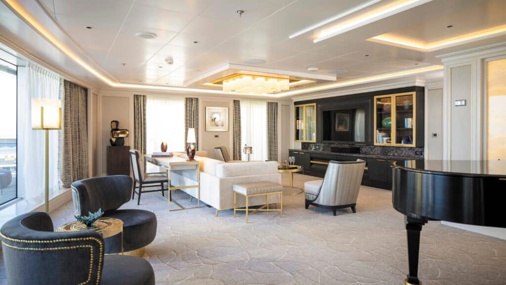 The image depicts the interior of a living room on a cruise ship. The room features typical cruise ship decor, including comfortable seating arrangements, a coffee table, and a small entertainment area.