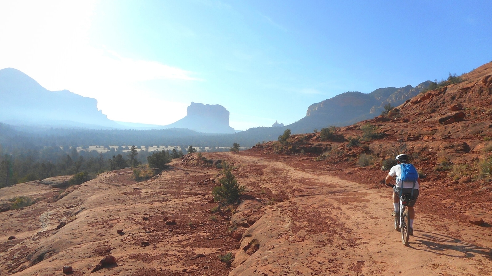 A photograph capturing a cyclist riding through arid terrain in Sedona, Arizona, with mountain cliffs in the background. The cyclist is depicted navigating the rugged landscape, showcasing the region's natural features and challenging terrain.