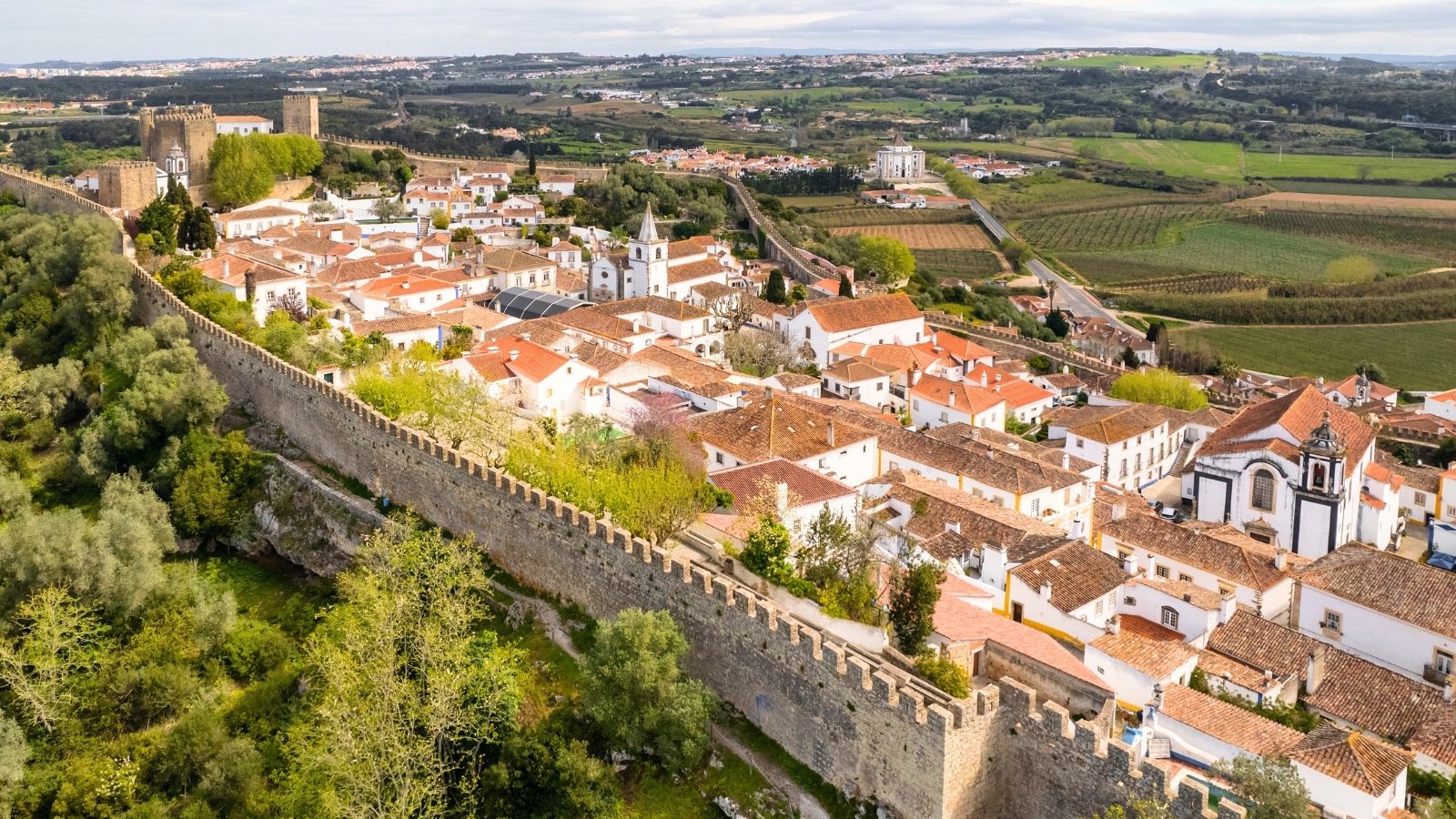 An aerial view capturing the historic walled town of Obidos near Peniche, Portugal. The image provides a drone perspective of the medieval fortress and historic castle within the town. The architectural elements of the walled town, including the castle, are prominent in the shot, showcasing the medieval features of Obidos.