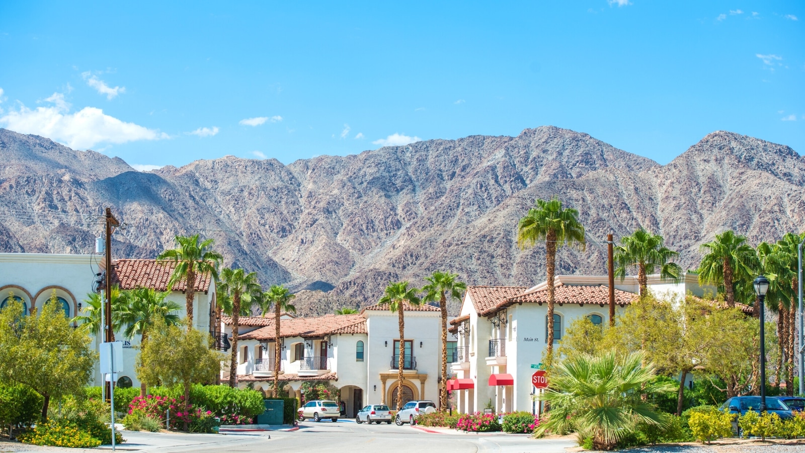 An image capturing the scene of Palm Springs' Old Town La Quinta. The photograph features two-story buildings surrounded by palm trees and abundant greenery. In the background, mountains provide a scenic backdrop against the clear sky.