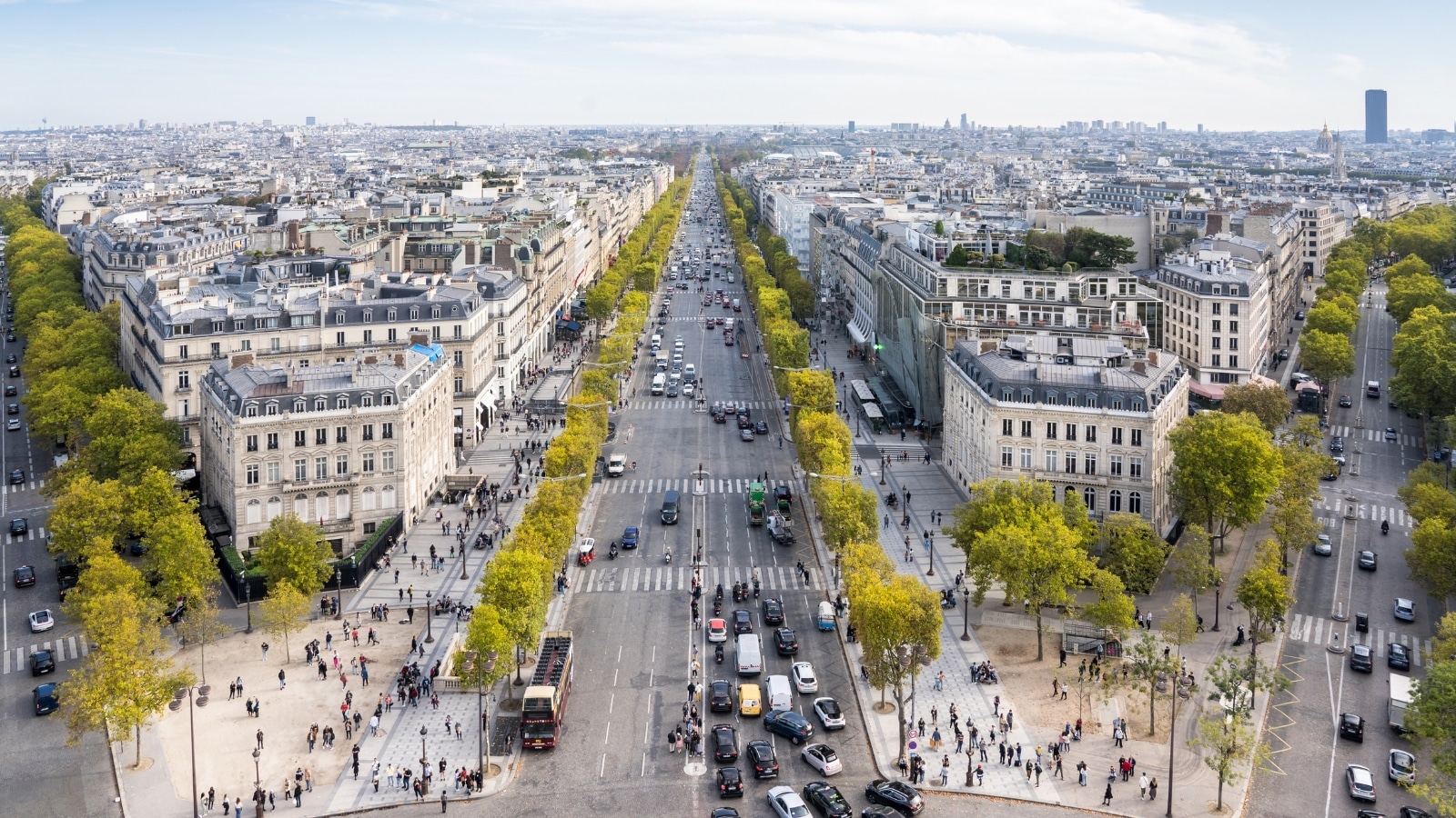 A panoramic view showcasing Paris from the Arc de Triomphe. The image reveals three traffic avenues extending in different directions, featuring wide car lanes with congested traffic, indicating potential traffic congestion. Numerous pedestrians are observed navigating the area. Alongside the roads, trees with green foliage contribute to the cityscape.