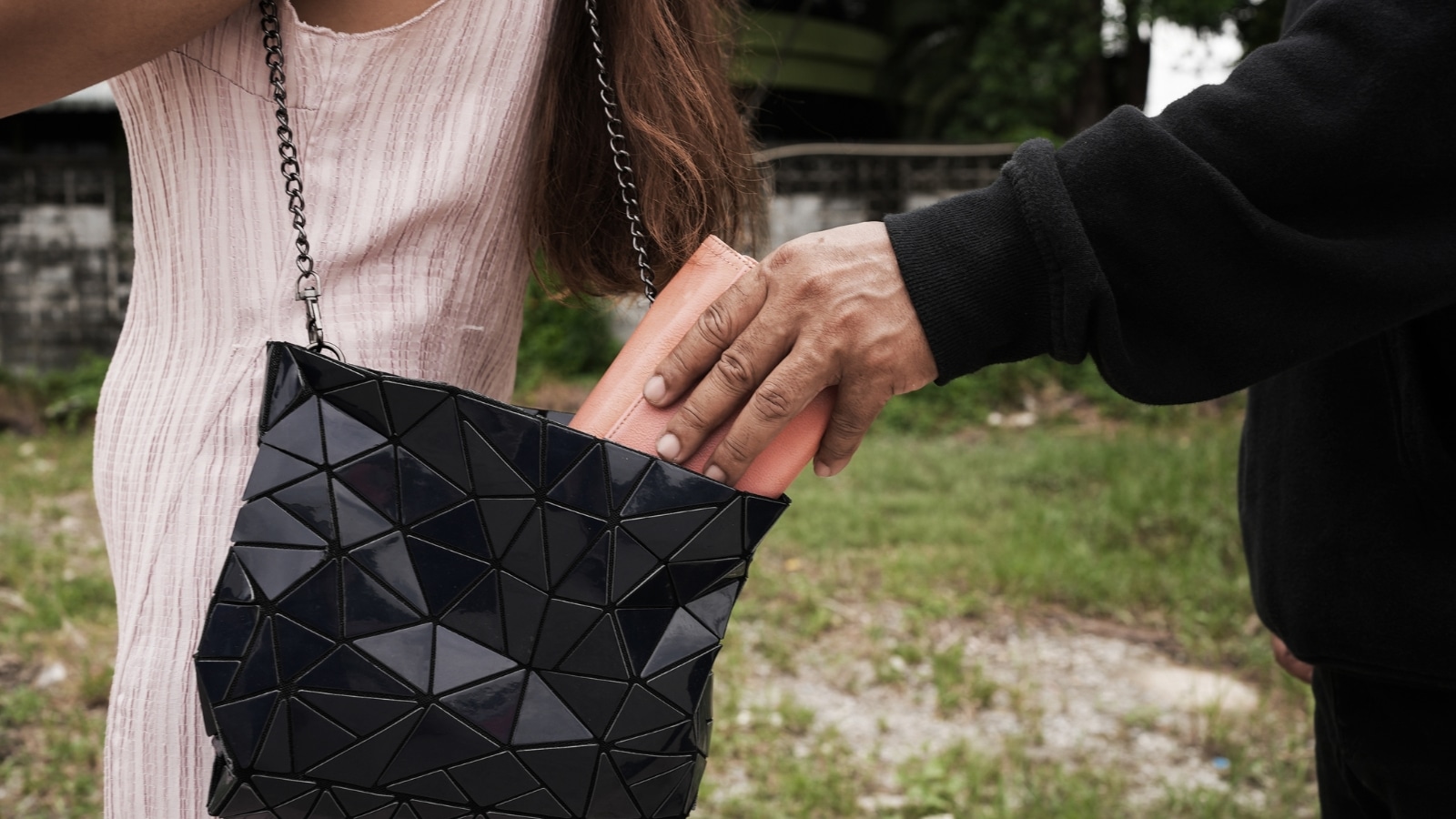  A close-up image capturing an incident of theft, with a man's hand discreetly taking an orange wallet from a woman in a pink dress, who is holding a handbag. 