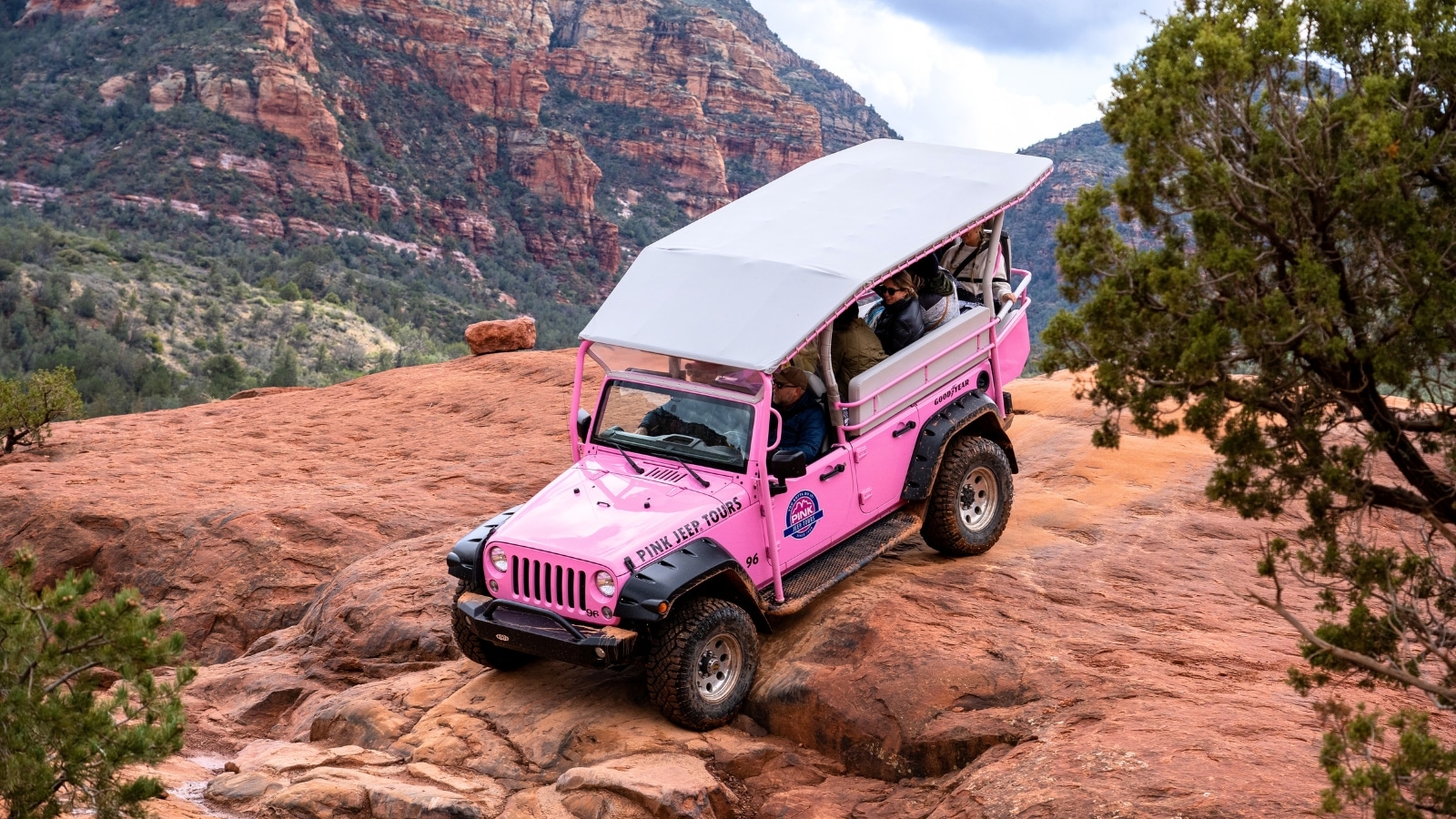 A pink SUV from Pink Jeep Tours traverses rocky terrain in Sedona, Arizona. The vehicle is occupied by passengers, visible through the windows. The rocky landscape and rugged terrain showcase the adventurous nature of the tour.