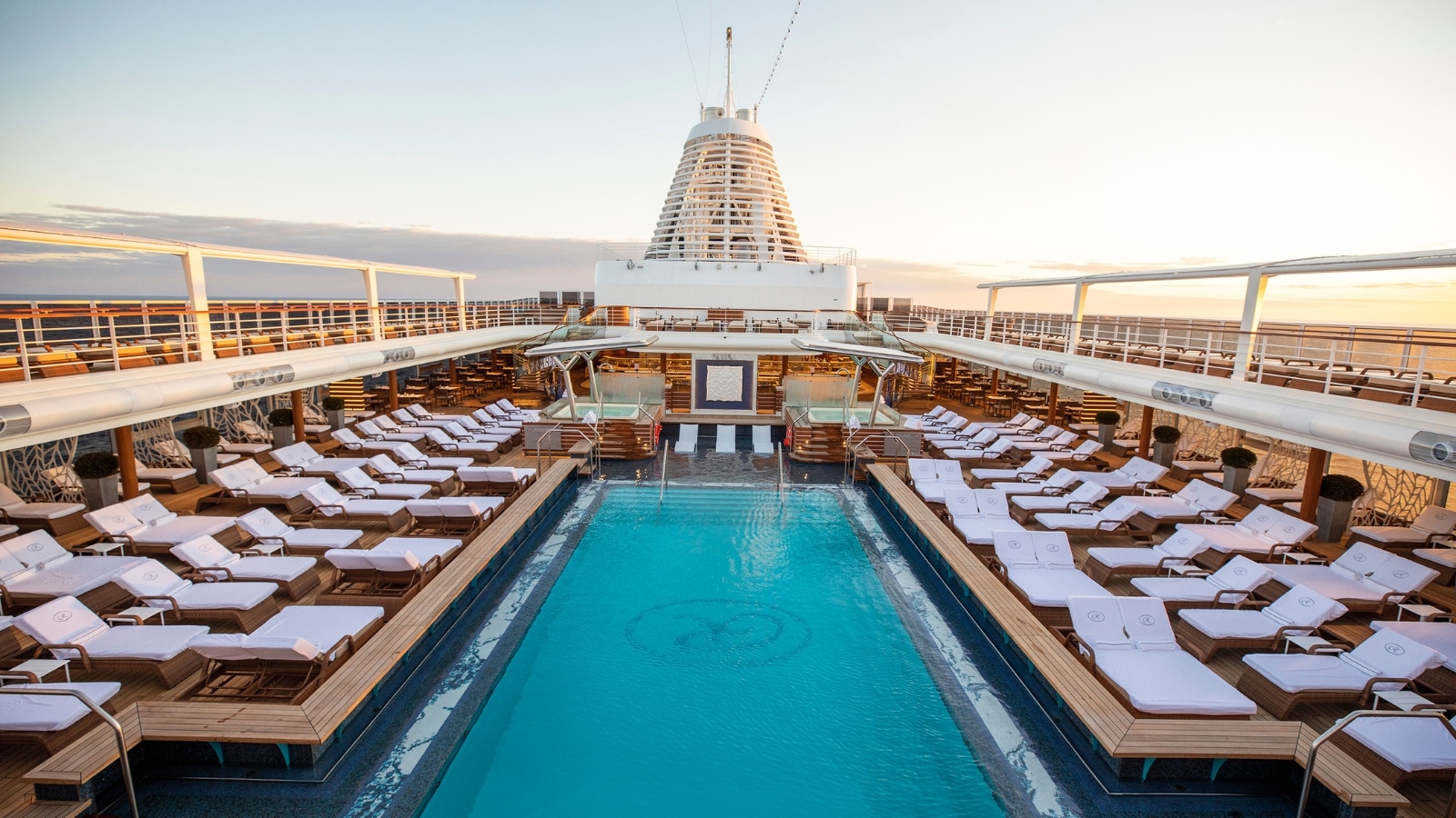 The image depicts a close-up view of the Pool Deck on a large ship, showcasing numerous white sun loungers arranged around the pool area.