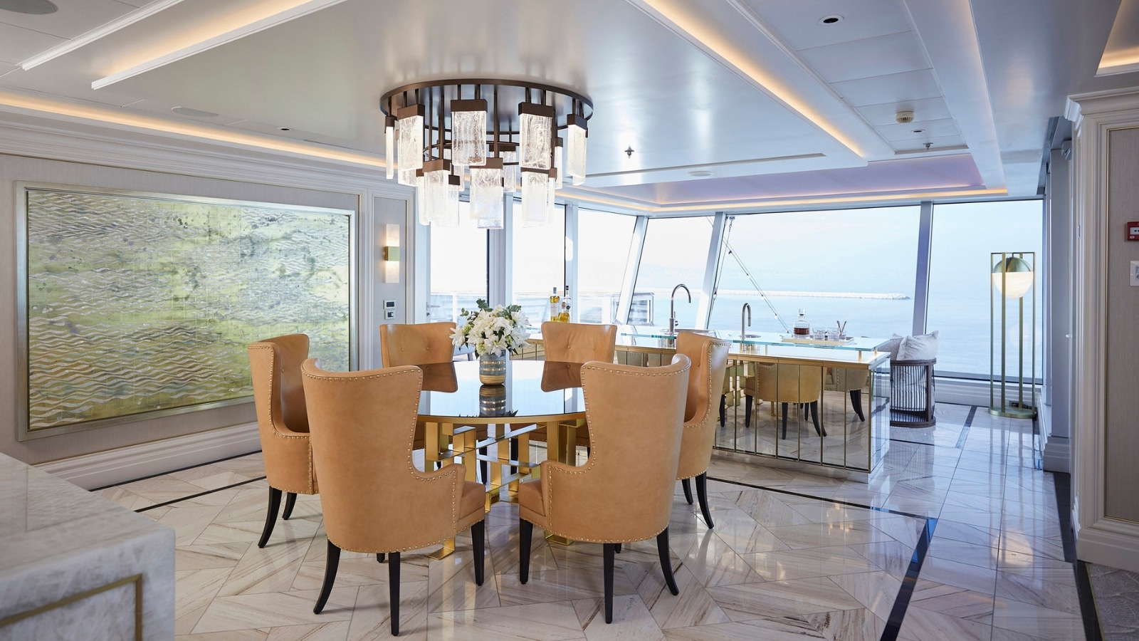 The image depicts the interior of a Regent Suite Dining Area. The room has a round dining table with soft beige armchairs. Marble floors, large panoramic windows, a large luxurious chandelier, a painting on the wall.