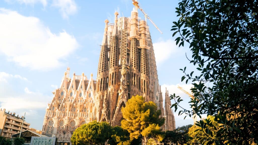 A photograph showcasing the exterior of Sagrada Familia. The image emphasizes the intricate architectural details of the basilica's facades, featuring distinct sculptural elements.