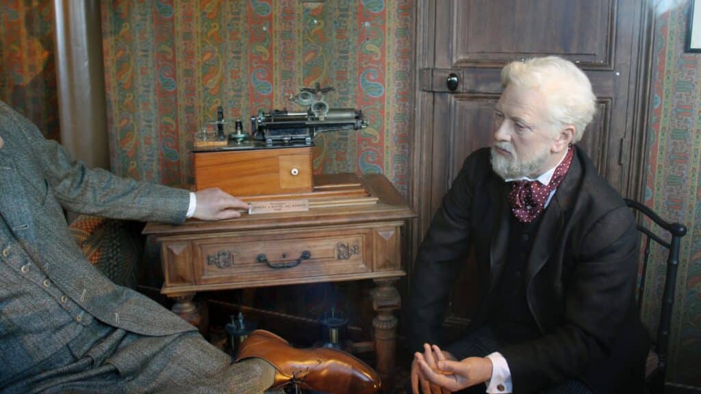 A photograph depicting wax figures of Eiffel and Edison in the Eiffel Tower's secret room at the top. The image captures the interior setting with the wax figures placed in a historical context.