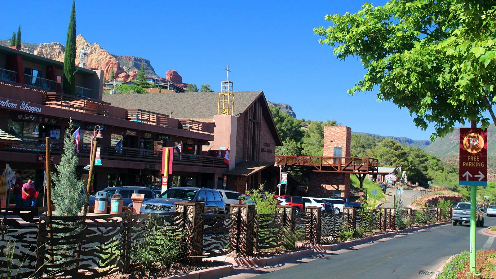 A photograph depicting a row of buildings in downtown Sedona, Arizona. Cars are parked along the street under the buildings, indicating urban activity.