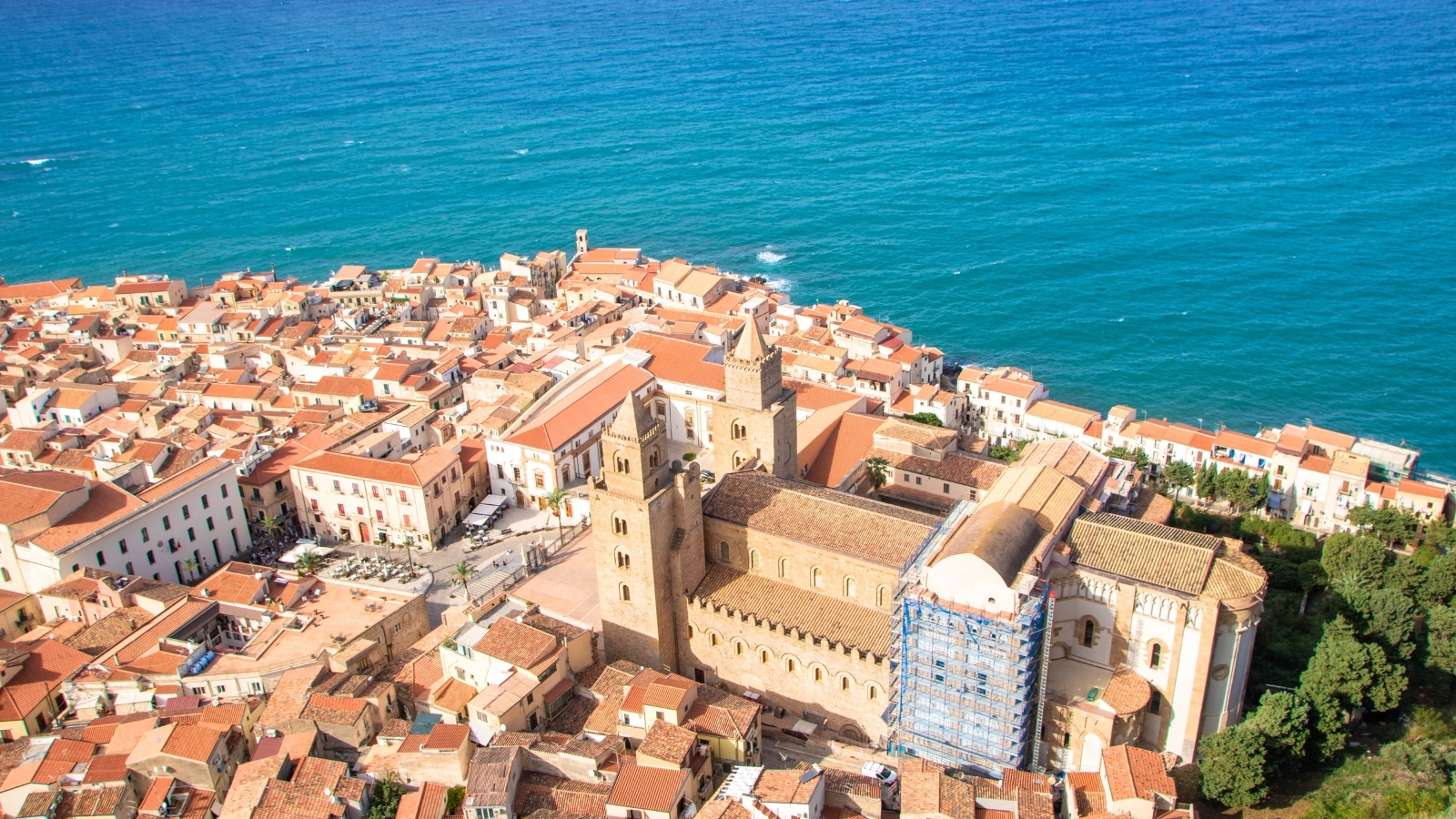 A photograph showcasing the town of Cefalu from the vantage point of La Rocca in Palermo province, Sicily, Italy. The image captures the expansive landscape with Cefalu nestled within it. The terrain features a combination of coastal areas, urban structures, and natural elements.