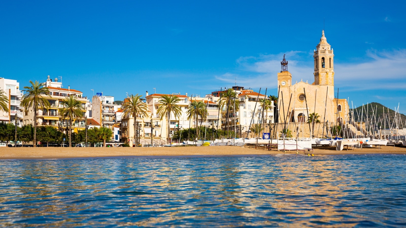A view from the sea capturing Sitges beach and the Church of St. Bartholomew and Santa Tecla on a sunny fall day in Catalonia, Spain. The image features the coastal landscape with the beach and prominent church in the background.