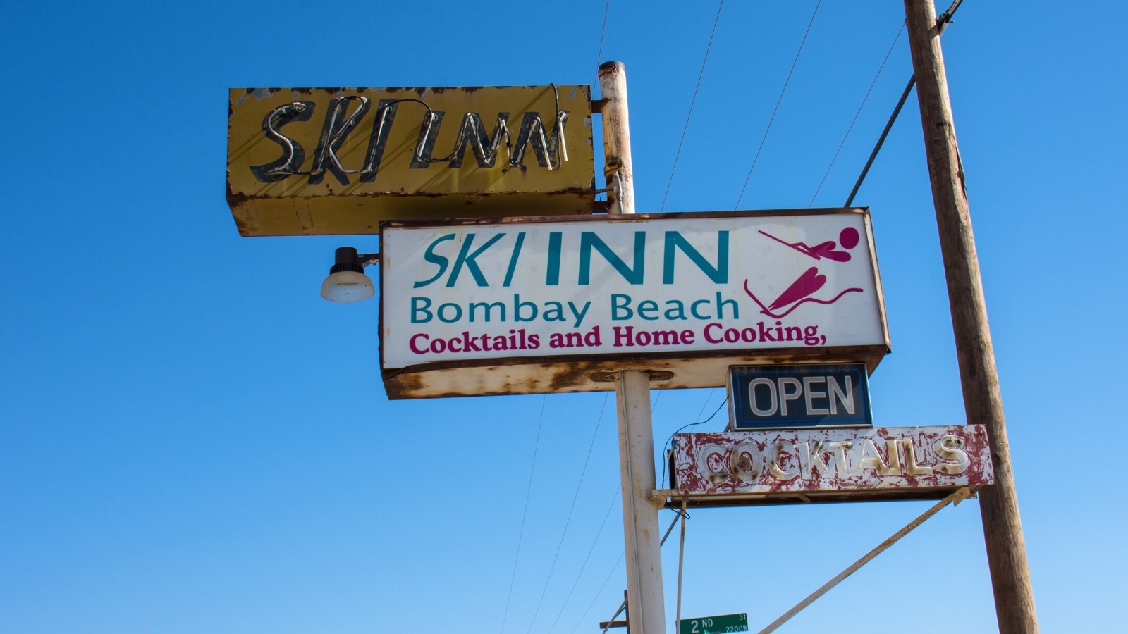 A photograph capturing several signs mounted on poles against a clear blue sky. The signs read 'Ski Inn,' 'Bombay Beach,' 'Cocktails,' and 'Home Cooking.'