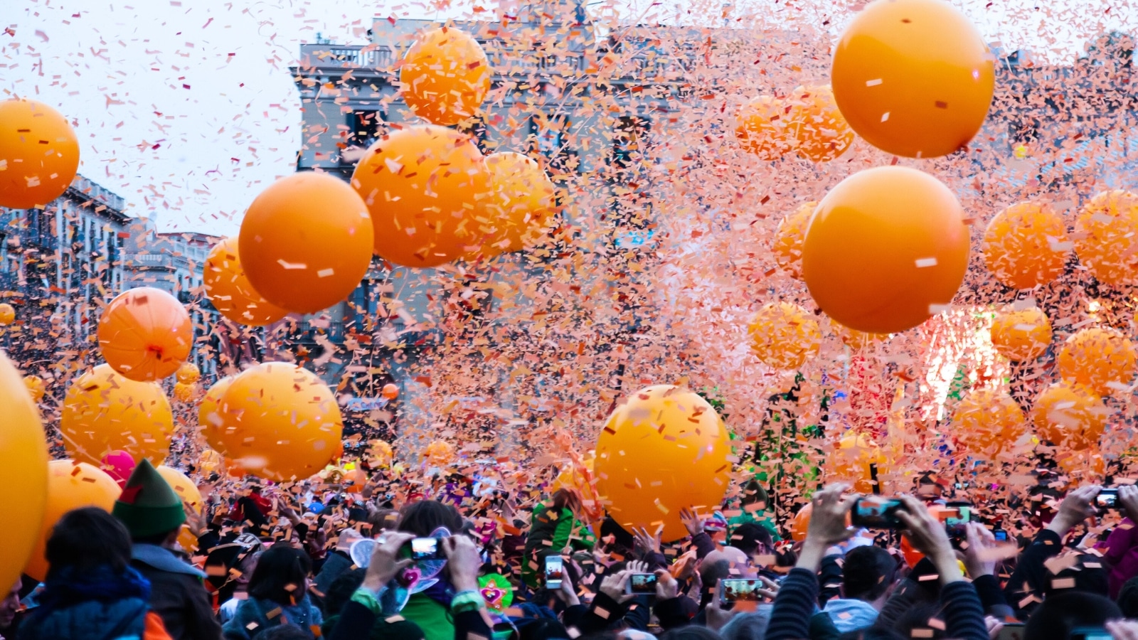 A photograph depicting the Spain Barcelona Carnival, featuring a crowd of people amidst a festive atmosphere. Numerous orange balloons are seen floating above the crowd, adding to the celebratory ambiance. Firecrackers and confetti are dispersed throughout the scene