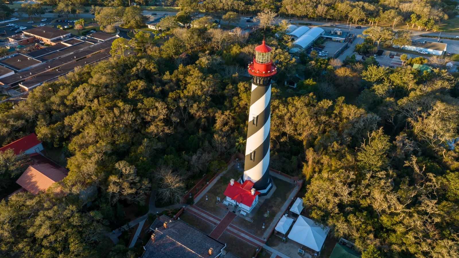 The image shows a top view of the St. Augustine Lighthouse, capturing its iconic structure from an elevated perspective. Surrounding the lighthouse are lush greenery and perhaps some nearby buildings or structures.