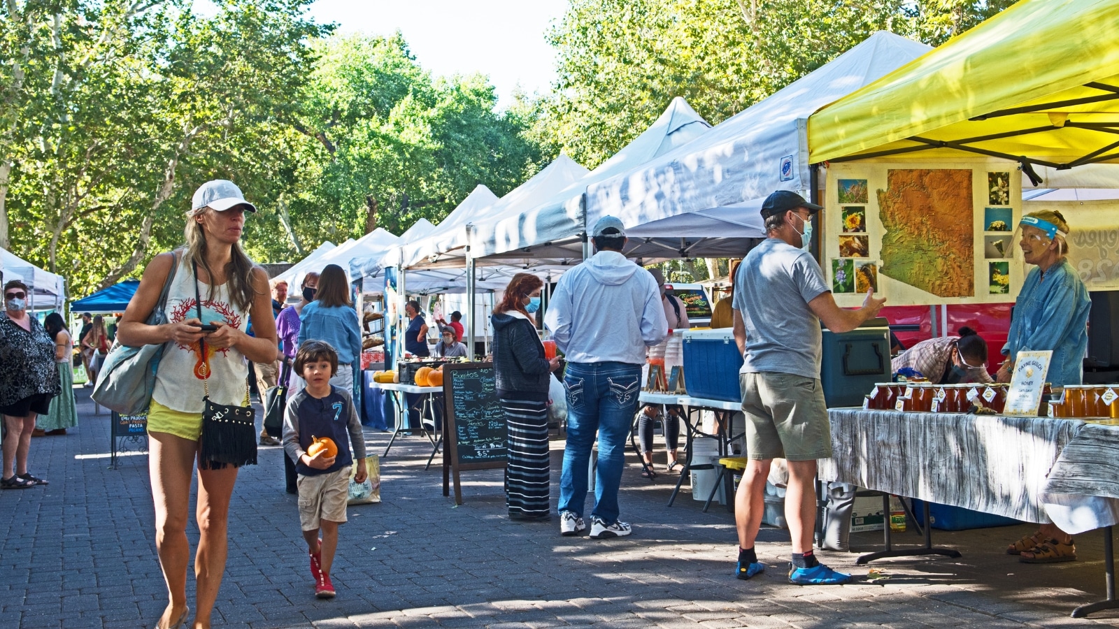 The image captures the scene at the St. Augustine Farmers Market, where people are observed walking along the stalls selling various goods.